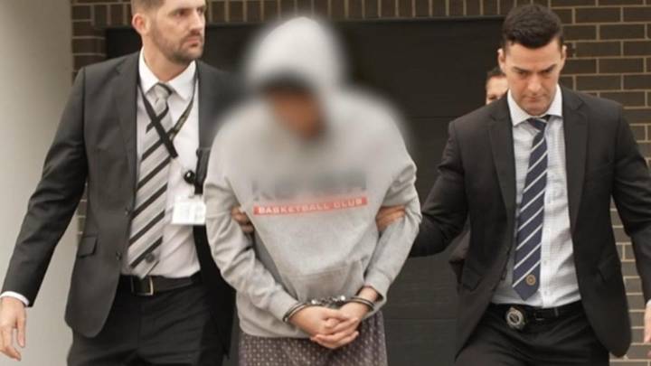 Man in hoodie escorted by two officials in business suits outside brick building