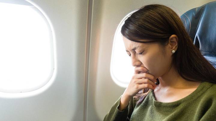 Woman in green sweater sits by airplane window, hand to face, appearing worried or upset