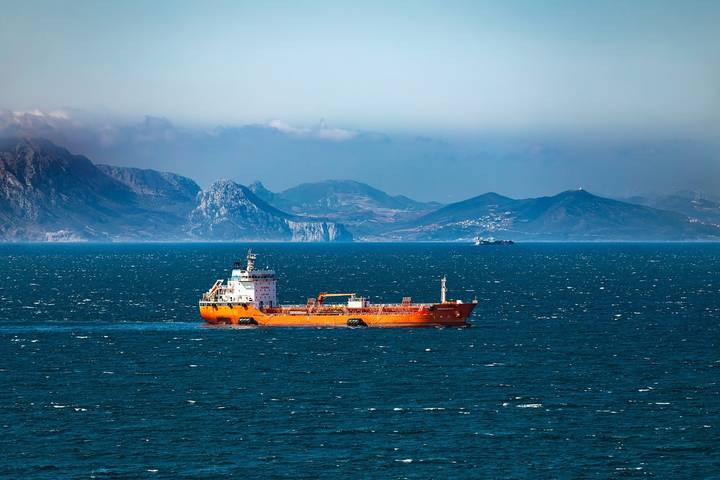 Tanker ship at sea with dramatic mountainous coastline in background, representing strategic shipping routes.