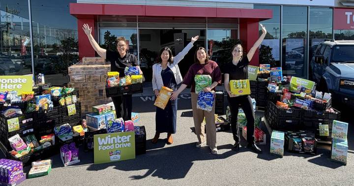 Four volunteers holding food items celebrate outside a Toyota dealership surrounded by stacked boxes of collected food for Winter Food Drive