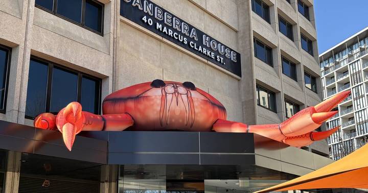 Giant red inflatable crab sculpture with extended arms mounted on Canberra House building facade