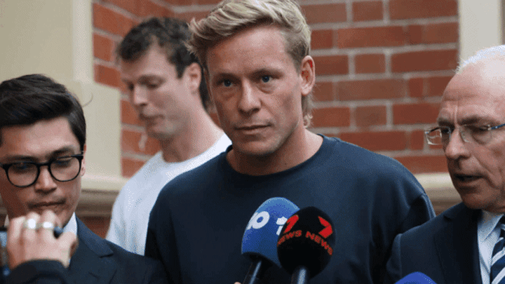 A man addresses multiple microphones at a press event; other men visible beside and behind him against a brick wall.