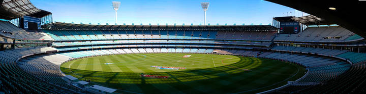 Large football oval with blue seating, green grass field, viewed from grandstand during daytime.