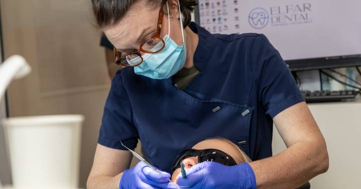 Dentist in scrubs, mask and gloves examines patient's teeth with dental instrument in clinical setting.