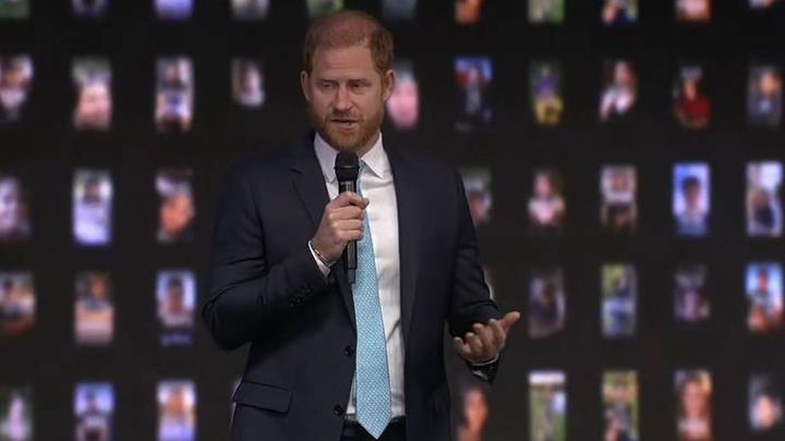 Man in dark suit and blue tie speaking at microphone, blurred photos on wall behind