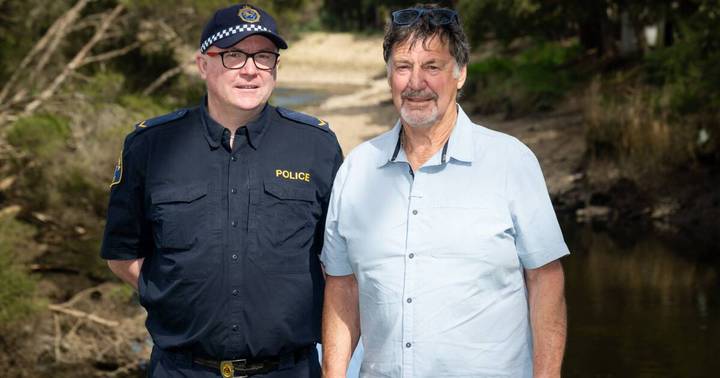 Police officer in uniform standing with older man in light shirt against rural background
