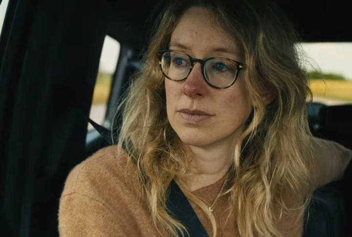 Nathan Fielder. Woman wearing glasses and tan cardigan sitting in car, looking to side with serious expression