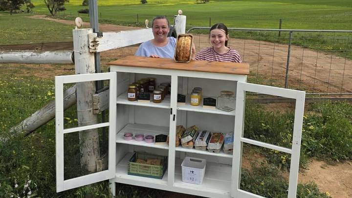 Two people at white cabinet roadside pantry stand with jars, goods and baked bread displayed