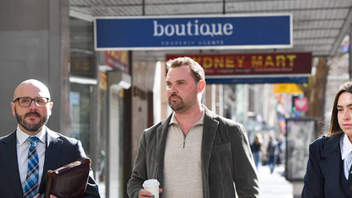 Two men walking on Australian city street past boutique storefront, one in suit with briefcase, one in casual cardigan with coffee