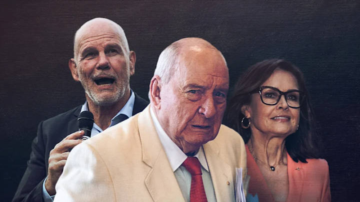 Three people at public event: man speaking with microphone, older man in beige jacket, woman in glasses and coral top