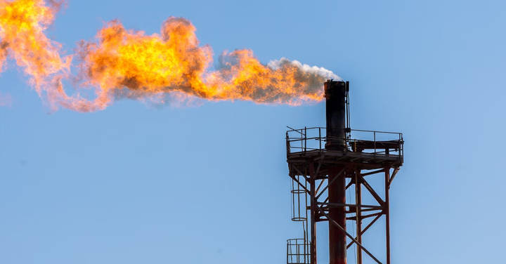 Tall industrial flare stack emitting dramatic flames and fire against clear blue sky