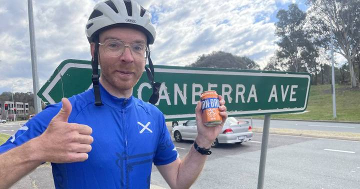 Connor McElwaine. Cyclist in blue jersey and helmet holding jar in front of Canberra Avenue sign