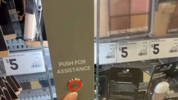 Hand pointing at red assistance button on locked glass cabinet containing $5 makeup palette in retail display