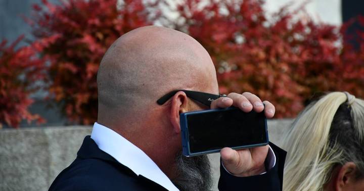 Tyson Bonfield. Bald man in business attire holding device to face, autumn foliage background