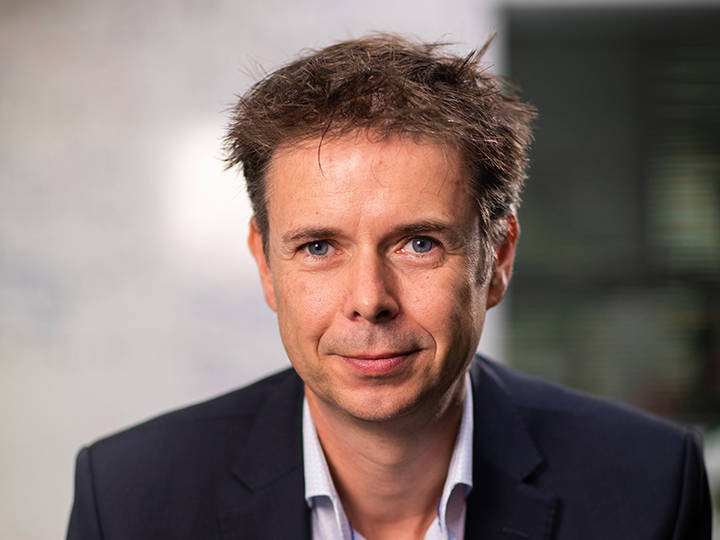 Professional headshot of man in dark blazer and white shirt, blurred background