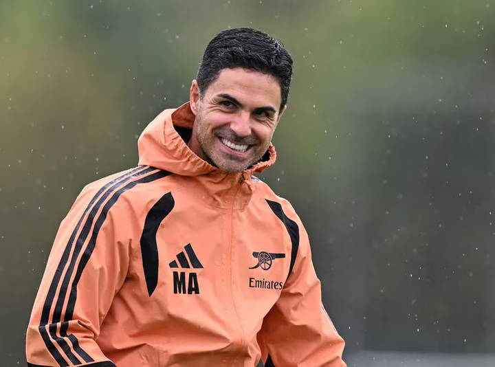 Smiling man in coral Arsenal Adidas training jacket with Emirates branding, outdoors in rain.
