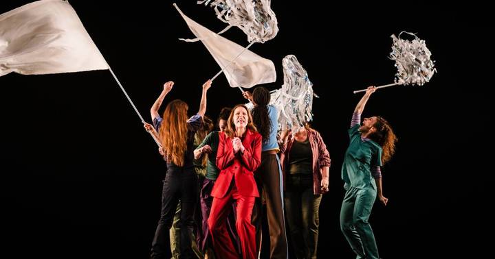 Performers on stage in theatrical production wearing varied costumes, waving translucent props