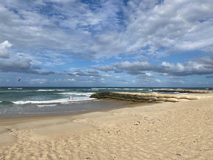 File image: Sandy beach with waves and rock formations under blue sky with clouds.