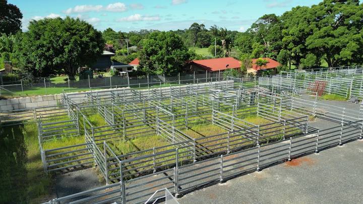 Aerial view of cattle yards with metal pens and red-roofed farm buildings in rural landscape.