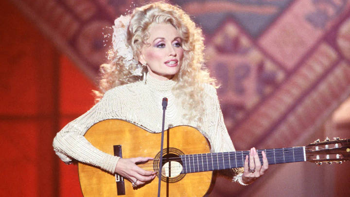 Dolly Parton. Woman with blonde curled hair in white sweater performing with acoustic guitar on stage