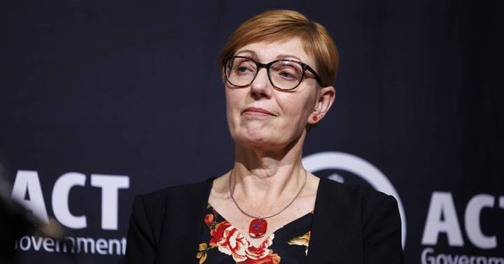 Woman in glasses and black floral-patterned top speaking at public event against dark background