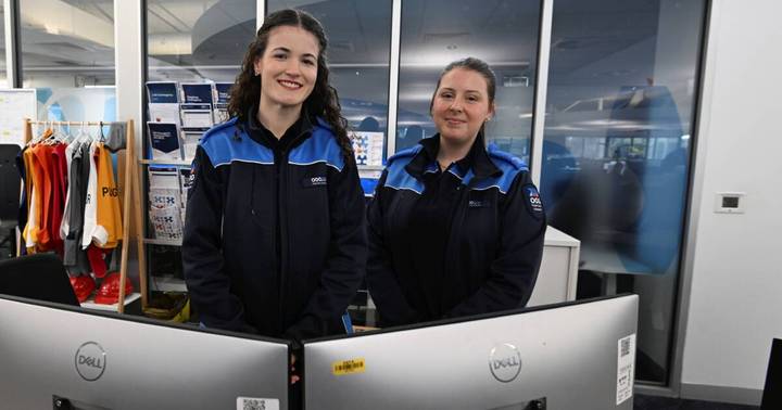 Two emergency services operators wearing 000 branded jackets at computer desks in a call center, with city views behind