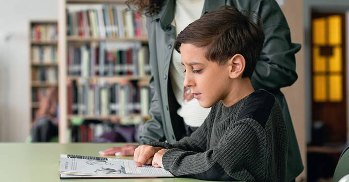 File image: Young student reading at table in school library with bookshelves visible, adult nearby.