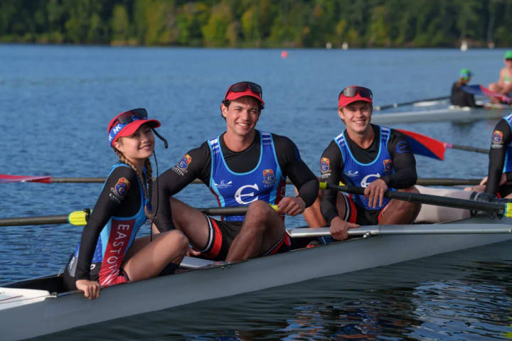 Three rowers in a racing shell on open water with forest background