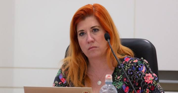 Woman with red hair in floral jacket speaking at microphone during official meeting