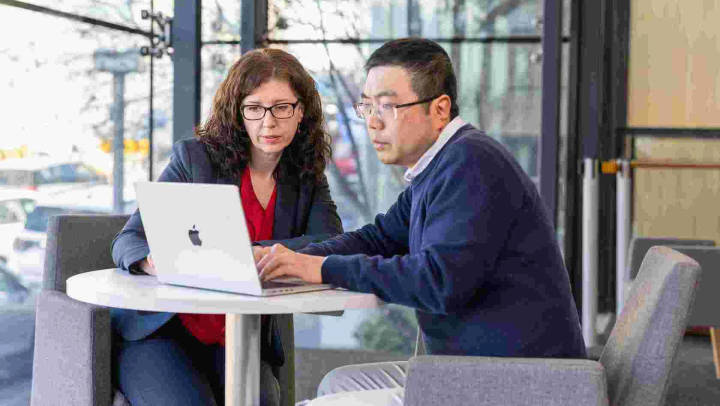 Two professionals reviewing laptop in modern office with city views behind