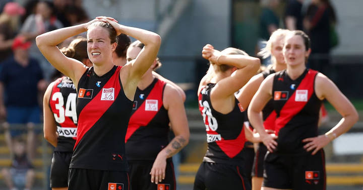Female AFL players in red-and-black Essendon uniforms on field, one with hand to head