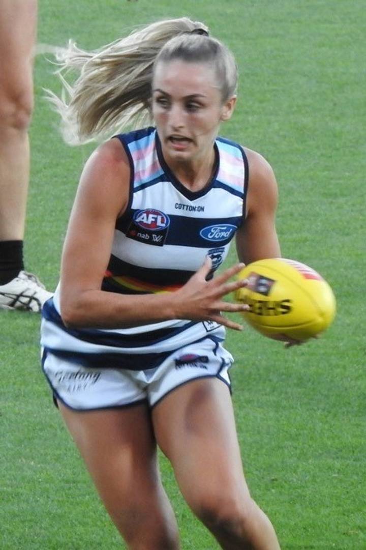 Amy McDonald. A female Australian rules footballer in a Geelong AFLW uniform runs with a yellow AFL ball clutched in both hands during a match.