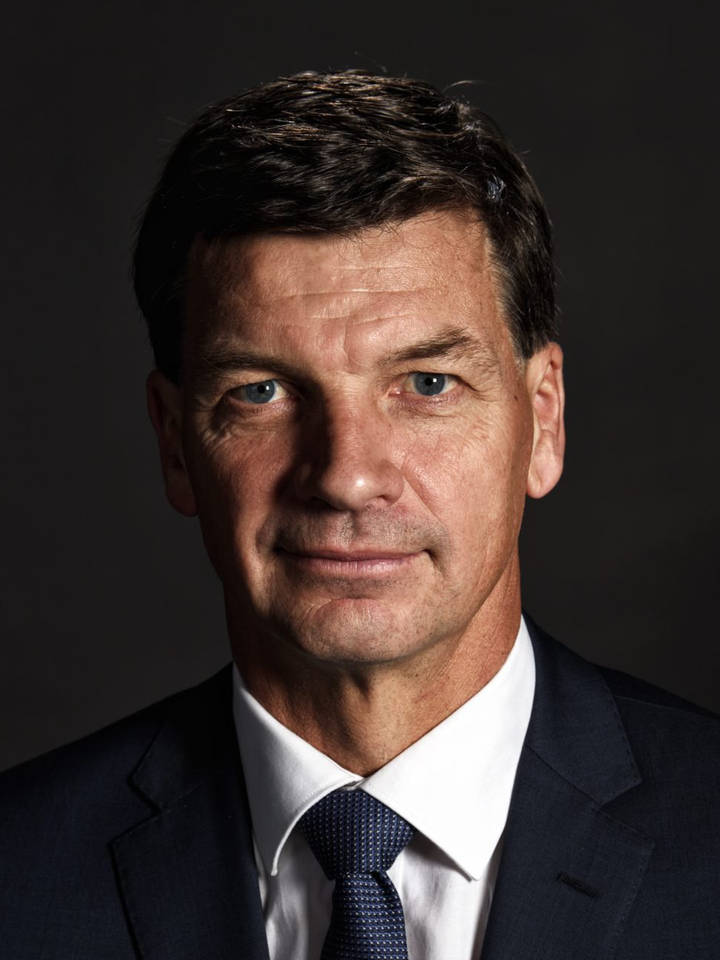 Angus Taylor. A close-up professional portrait of a middle-aged man with short dark hair and blue eyes, wearing a dark suit and white shirt, against a dark background.