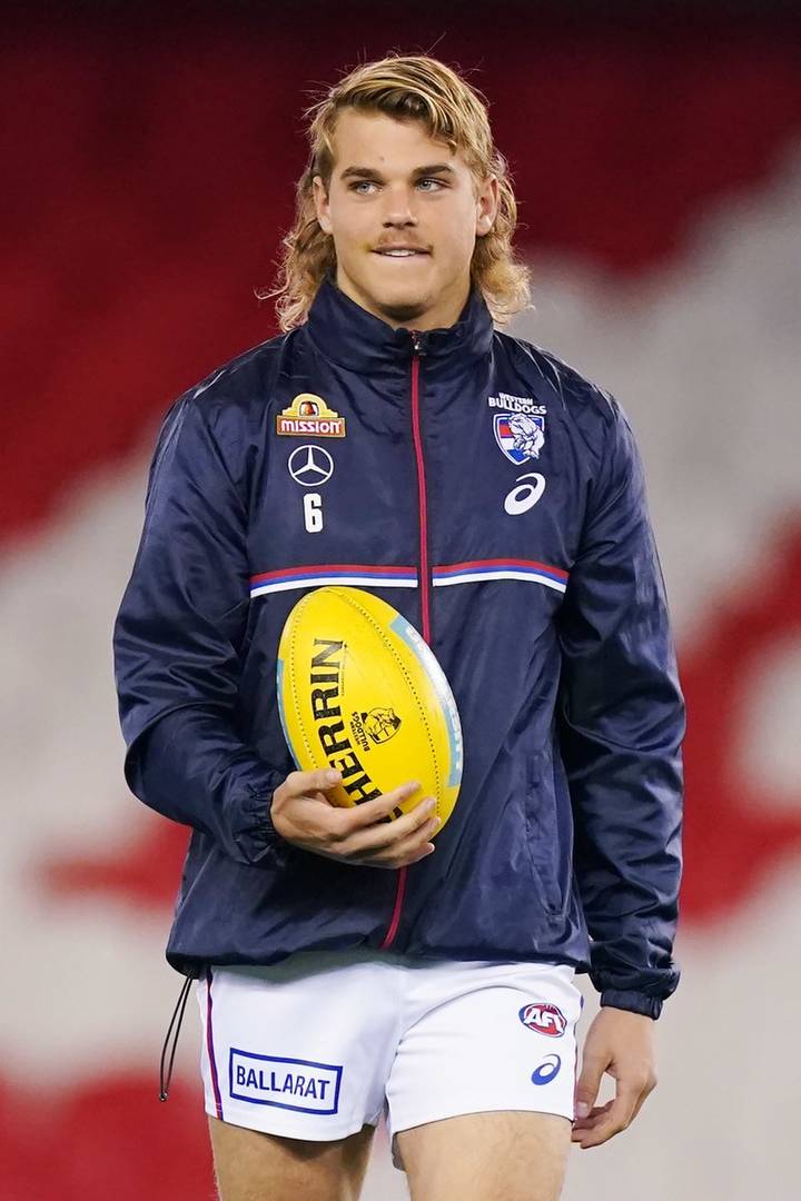 Bailey Smith. A young man with shoulder-length blonde hair wearing a navy Western Bulldogs AFL training jacket bearing the number 6, holding a yellow Sherrin football.