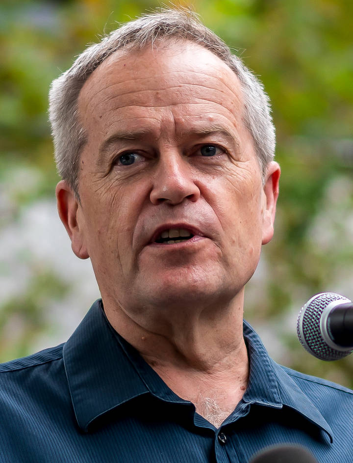A close-up photograph of a grey-haired middle-aged man speaking into a microphone outdoors with a blurred green background.