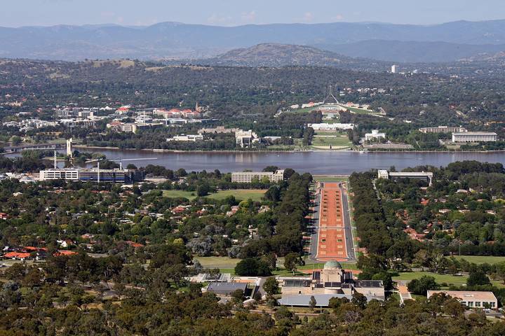 Aerial view of Canberra showing the Australian War Memorial and Anzac Parade leading to Lake Burley Griffin, with Parliament House visible on the far shore and mountains in the bac