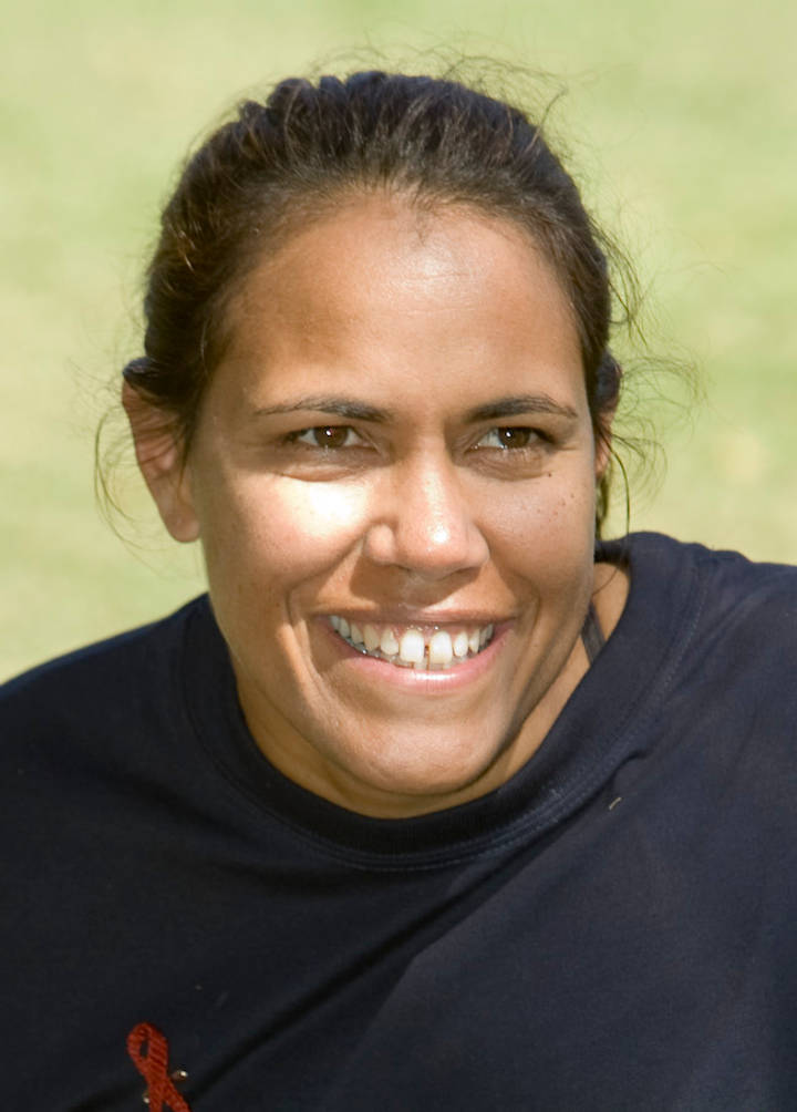 Cathy Freeman. A close-up photograph of a woman with dark hair pulled back, smiling broadly at the camera, wearing a dark top against a blurred green background.