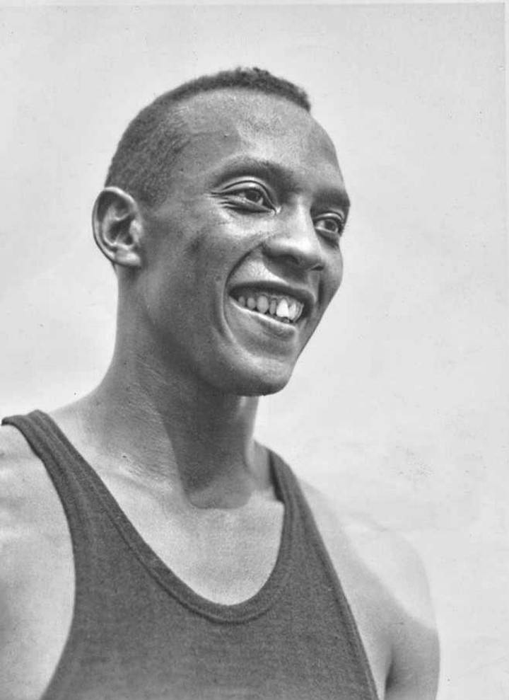 A black-and-white close-up photograph of a young man in an athletic tank top, grinning broadly, photographed from the chest up against a plain pale background.