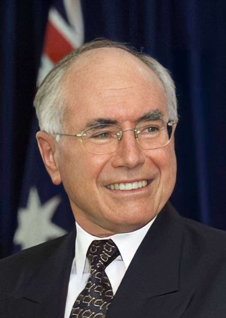 John Howard. A close-up portrait of an older man with white hair and wire-rimmed glasses, wearing a dark suit and smiling broadly against a blue background with a partial flag visible.