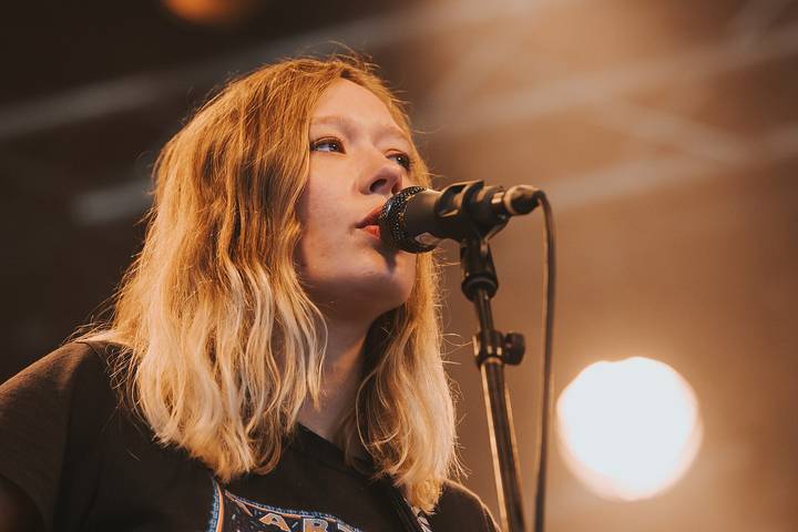 Julia Jacklin. A woman with shoulder-length blonde hair sings into a black microphone on a concert stage bathed in warm amber light.
