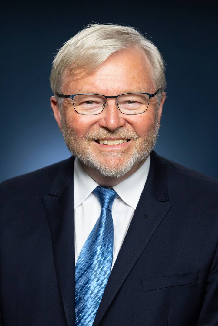 Kevin Rudd. A close-up professional portrait of an older man with white-grey hair and a short grey beard, wearing dark-framed rectangular glasses and a dark suit with a white shirt, smiling br
