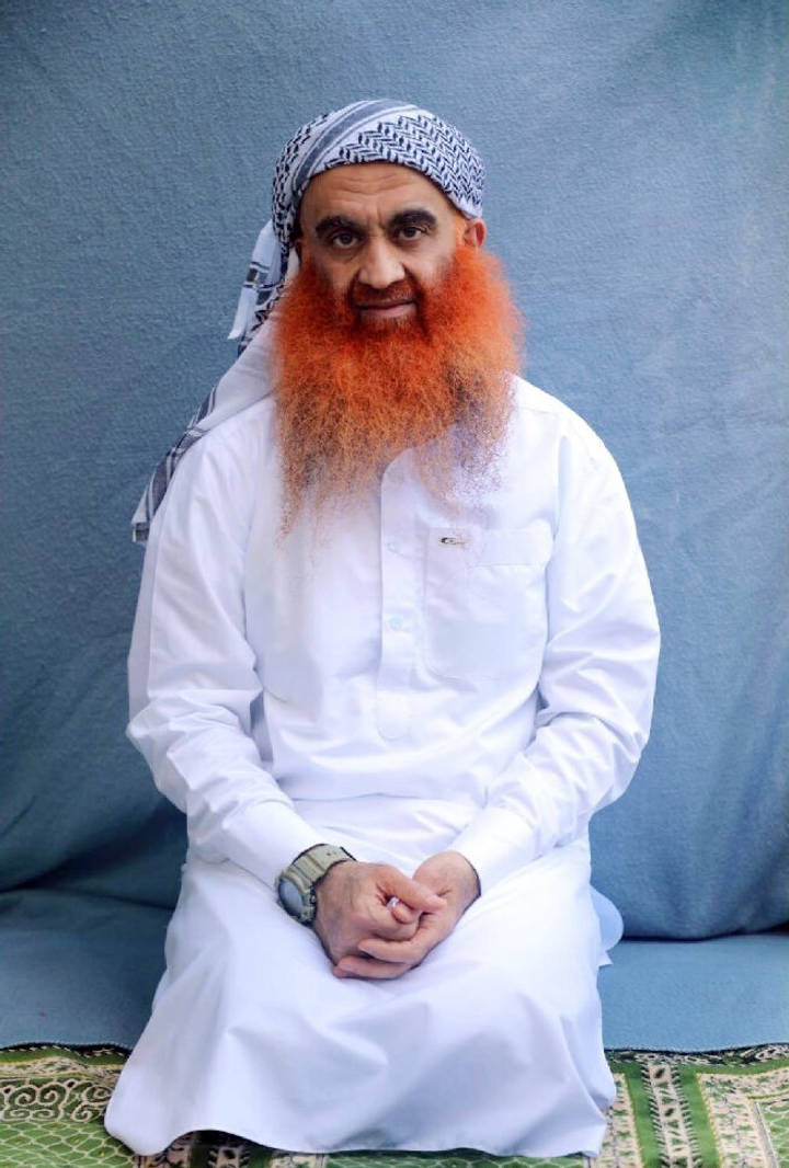 Khalid Sheikh Mohammed. A man with a long orange-dyed beard, wearing a white thobe and a black-and-white keffiyeh, sits calmly with his hands folded in his lap against a blue fabric backdrop.