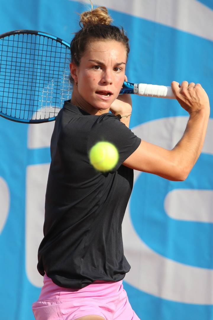 A female tennis player wearing a black shirt and pink shorts winds up for a backhand stroke, with a yellow tennis ball in the foreground and a blue banner in the background.