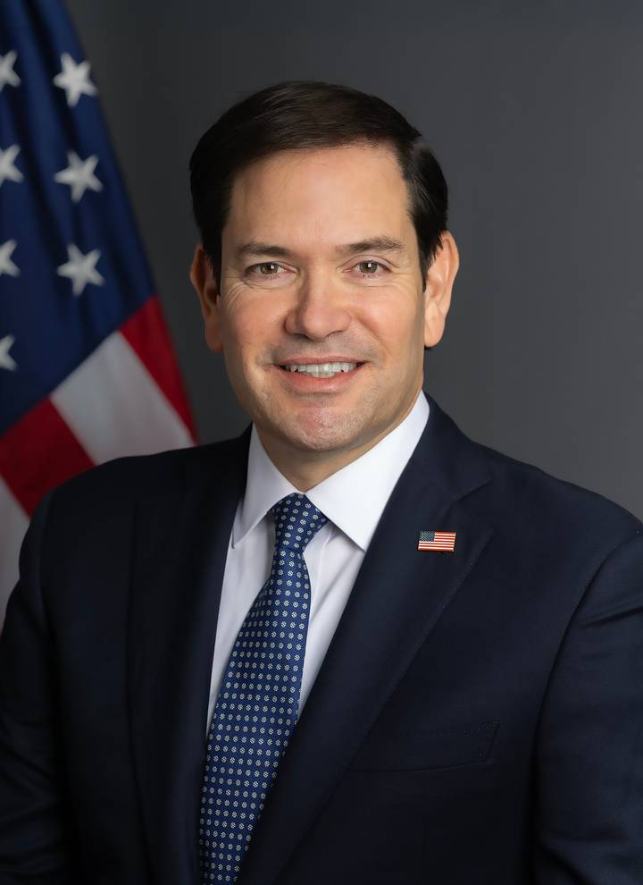 Marco Rubio. A man in a dark navy suit with a patterned blue tie and an American flag lapel pin smiles at the camera against a grey background, with a US flag visible to his left.