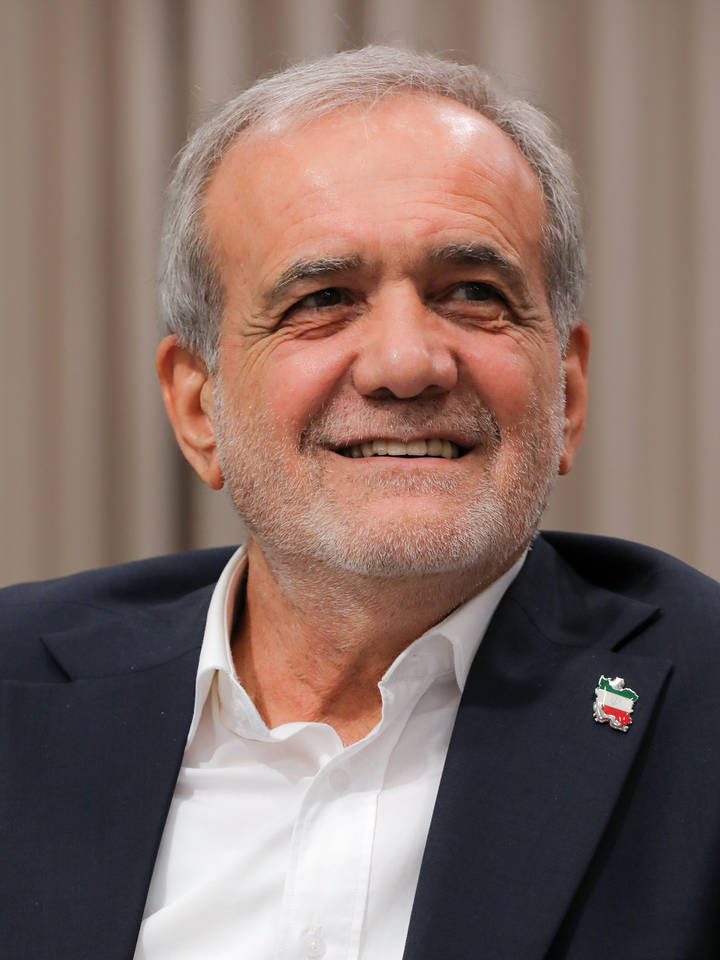 Masoud Pezeshkian. A grey-haired man with a short grey beard wearing a dark blazer over an open-collar white shirt and a small Iranian flag lapel pin, smiling broadly and looking slightly upward agai