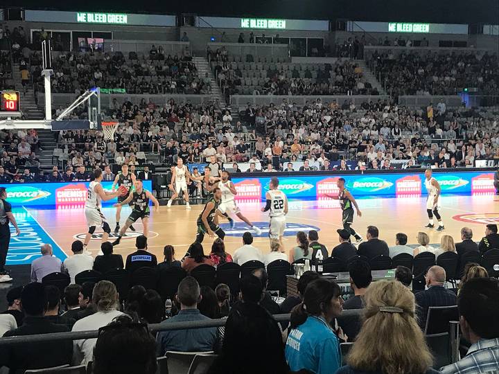 A professional basketball game in progress at a well-lit indoor arena, with players on the hardwood court, courtside advertising boards, and a large crowd filling the stands beneat