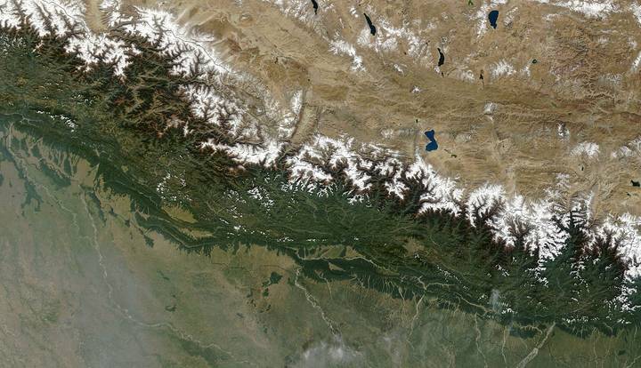 A satellite view of the Himalayan mountain range showing snow-capped peaks, green forested slopes, and the flat plains of the Indian subcontinent to the south with the Tibetan plat