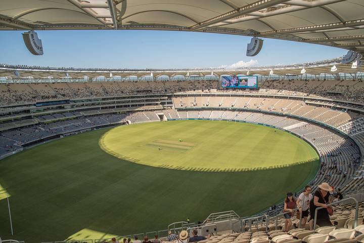 An elevated wide-angle view of Optus Stadium in Perth showing the full oval cricket ground with a prepared pitch, the distinctive tensile roof canopy, partially filled tiered stand