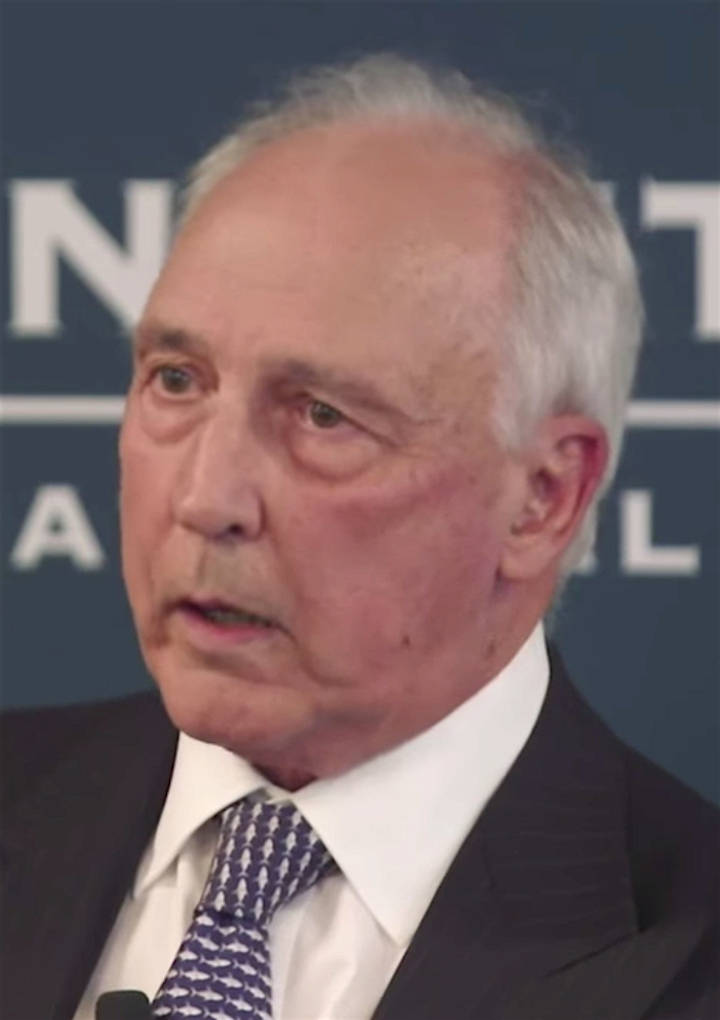 Close-up of an older man with grey hair wearing a dark suit and white shirt, speaking at what appears to be a press conference or official event.