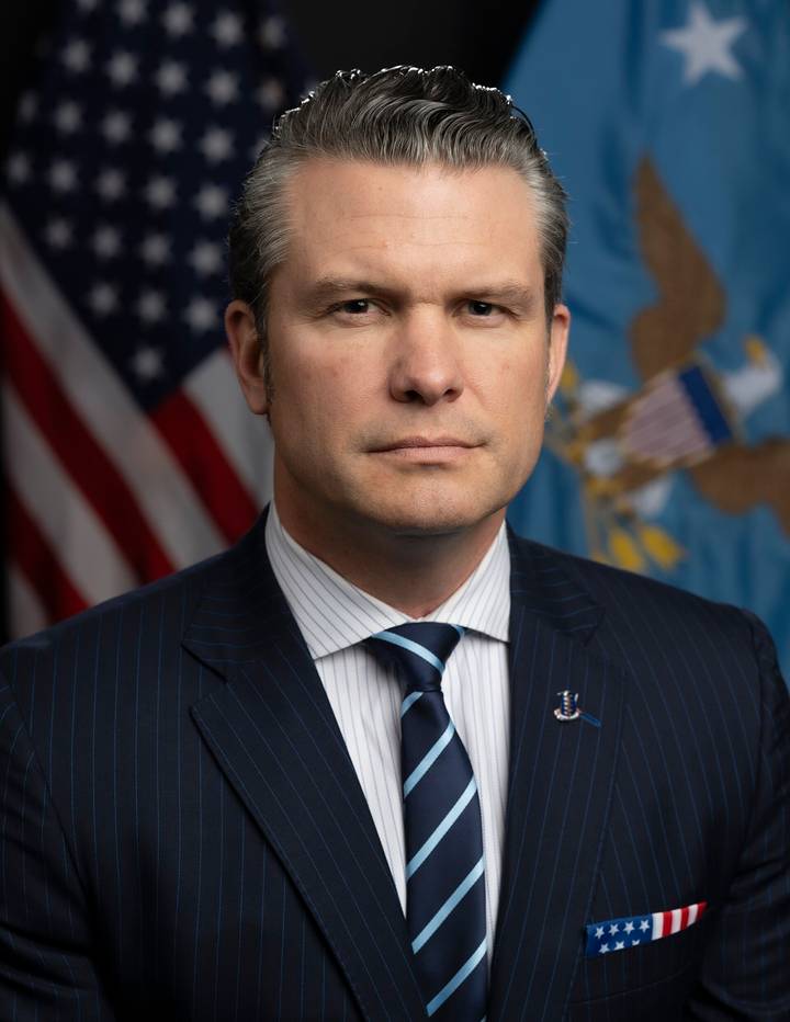 A man in a dark pinstripe suit and blue striped tie poses for a formal portrait in front of an American flag and a blue official government seal flag.